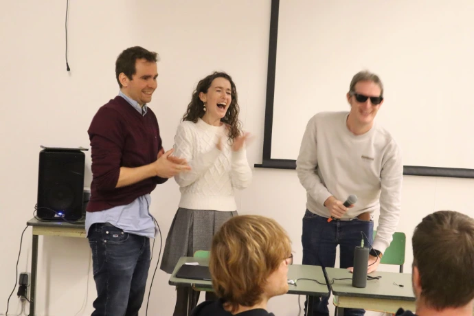 Three people are standing in front of a crow while applauding and laughing. One of them is holding a microphone