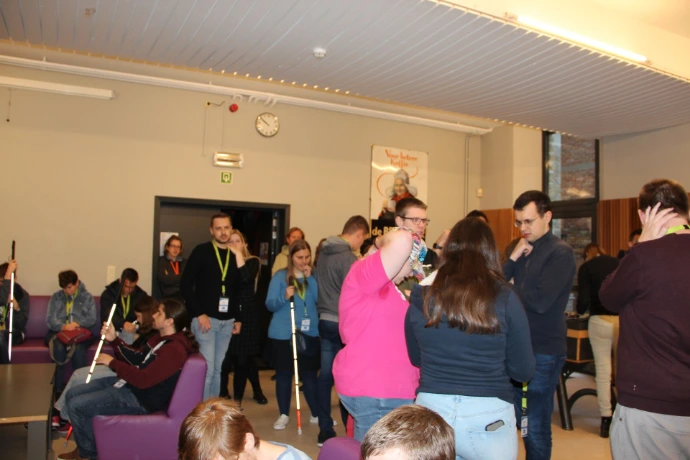 A large group of people is gathered in a mess hall. They are standing in groups and having conversations, some of them are using a white cane