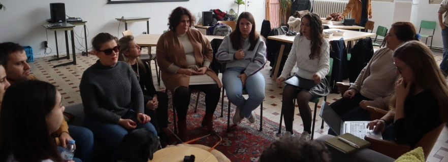 A group of young people sitting in a circular arrangement. There is a guide dog on the floor in the center, one person is taking notes on a note book.