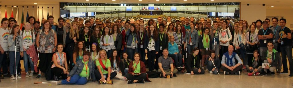 A group of over 100 people standing together at the EU Parliament and facing the camera. Several people have white canes, most people are smiling and some are giving a thumbs-up to the camera.