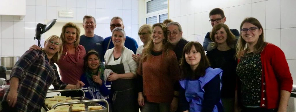 A group of 14 volunteers is standing in a kitchen, huddled up with arms on each others shoulders and smiling at the camera. Food that has been prepared is standing on a tray in front of them.